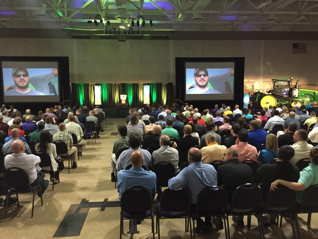 Strategic rollout venue setup — dual screens, John Deere equipment on stage, 1,800-seat convention center floor